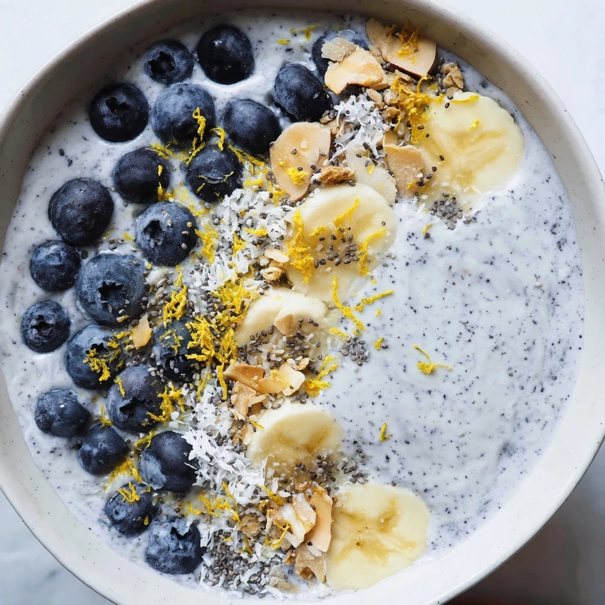 Bright blueberry and lemon smoothie bowl in a white bowl with crunchy granola and coconut flakes.
