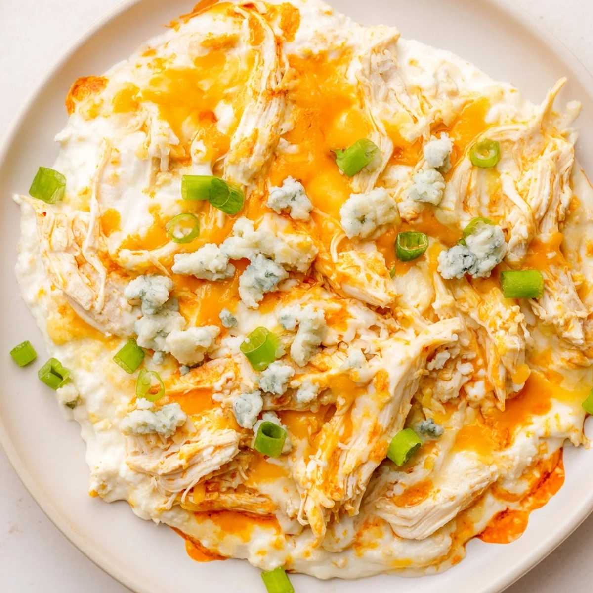 A close-up of creamy keto buffalo chicken dip bubbling in a white dish, garnished with fresh chopped green onions and crumbled blue cheese, perfect for low-carb game day snacking.
