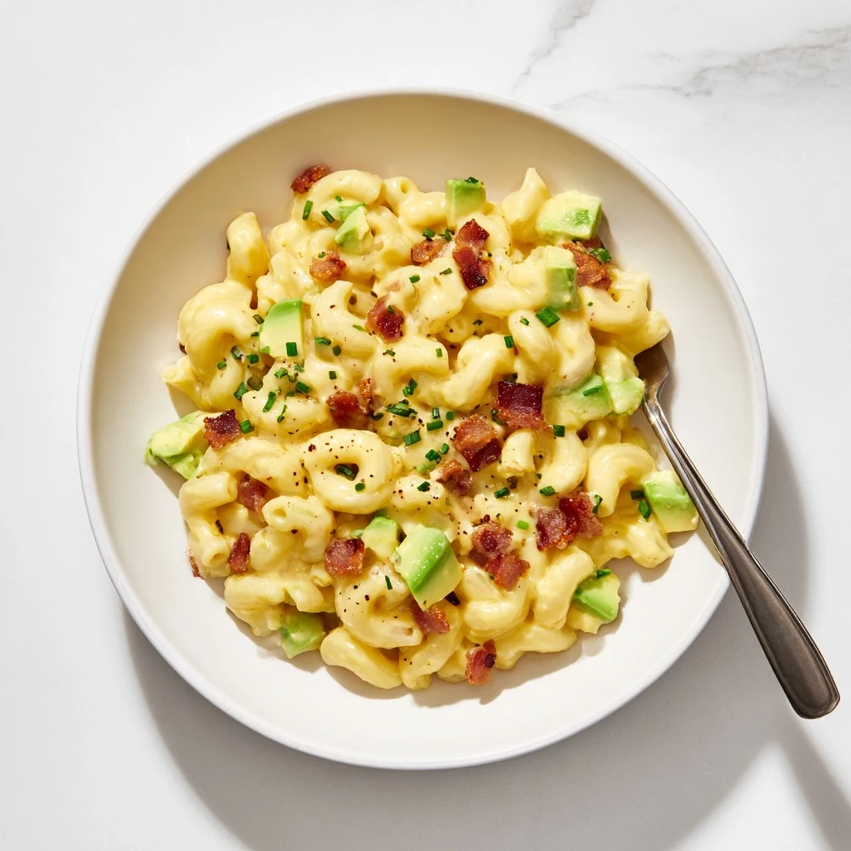 Homemade avocado and bacon mac and cheese with diced avocado and melted cheddar on a dinner plate.